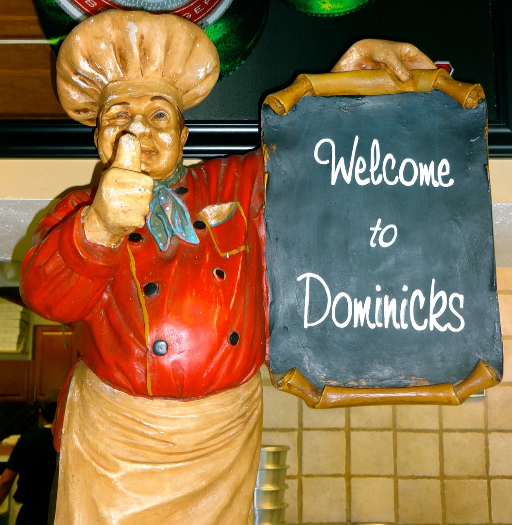 A statue of a chef holding a sign that says " welcome to dominicks ".